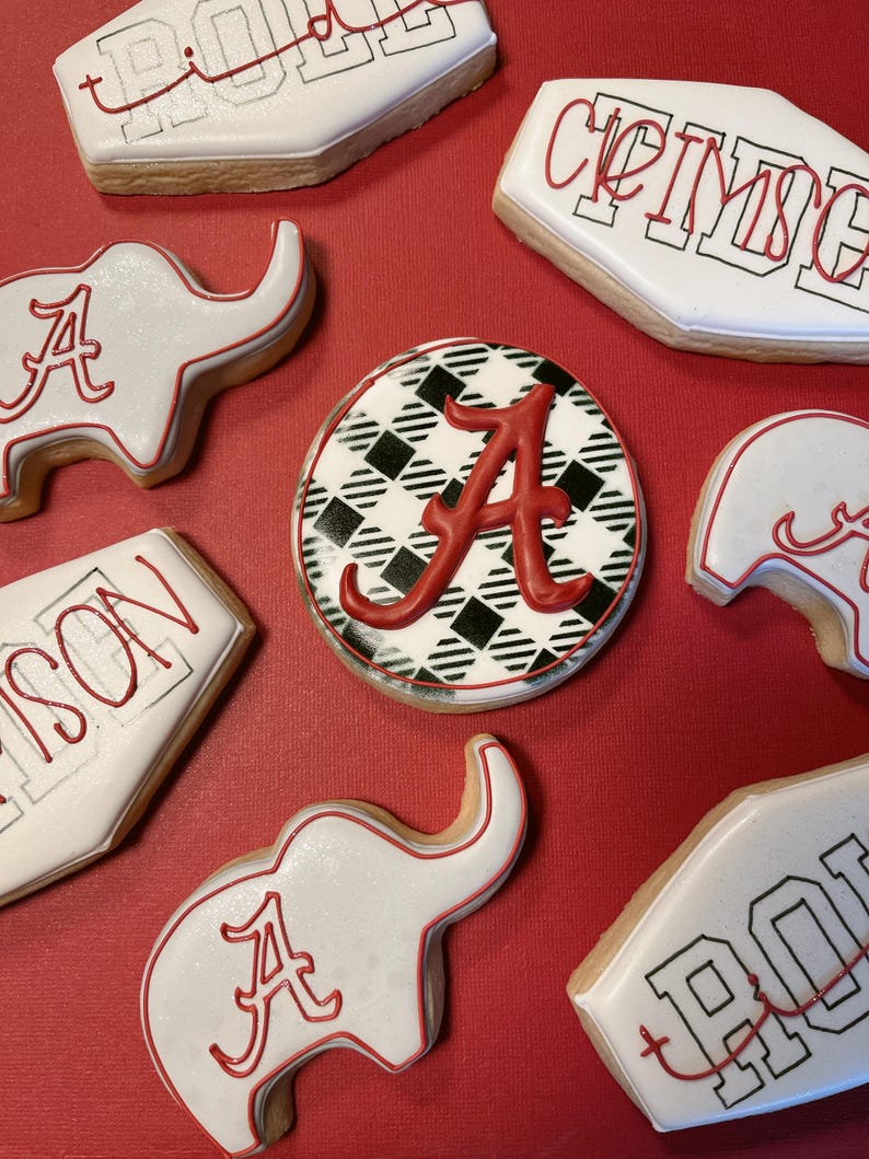 May include: Assortment of decorated sugar cookies on a red surface. The cookies feature white icing with red accents, including the letter "A" and team names. Some cookies are elephant-shaped, while others are rectangular or round.