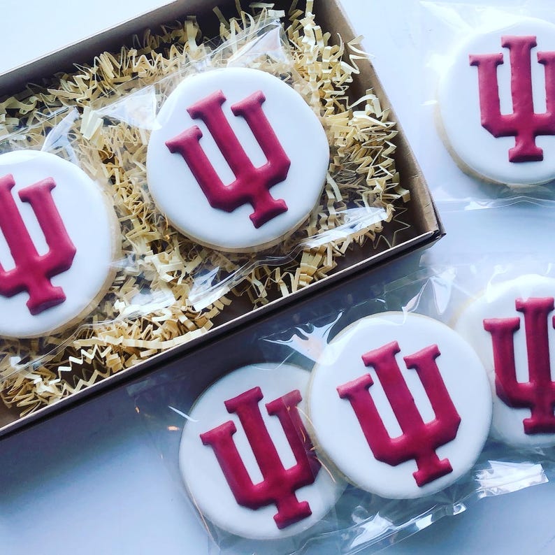 May include: Round sugar cookies with white icing and a maroon logo are packaged in a cardboard box with shredded paper. Several cookies are individually wrapped in clear plastic. The logo is a stylized letter "I".