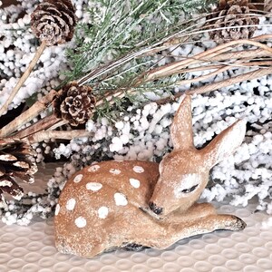 May include: A ceramic figurine of a fawn with white spots, lying down with its head resting on its front legs. The fawn is light brown with white spots. The background includes pinecones, branches, and faux snow, suggesting a winter theme.