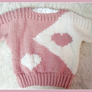 Puede incluir: Un suéter de punto rosa y blanco con un diseño de corazón. El suéter tiene mangas cortas y cuello redondo.