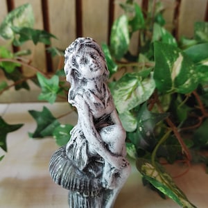 May include: A white and gray painted figurine of a fairy sitting on a mushroom. The fairy has long hair and is wearing a dress. The mushroom is detailed with ridges and has a dark gray base.