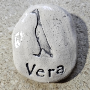 May include: A smooth, off-white, oval-shaped stone with a glossy finish. The surface features a dark, etched silhouette of a standing bird and the name Vera engraved in capital and lowercase letters below the bird figure in a dark color.