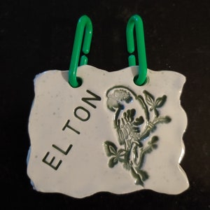 May include: White ceramic birdhouse with green bird and leaves design. The birdhouse has two green plastic hooks and the name "ELTON" written on the front.
