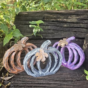 May include: Three decorative, open-design pumpkins in shades of orange, gray, and purple. Each pumpkin has a stem and a leaf accent. The pumpkins are displayed on a weathered wooden surface, suggesting a fall or harvest theme.