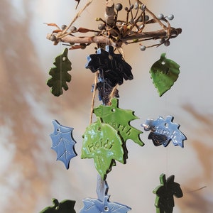 May include: A ceramic wind chime with green and blue leaves, each with a name written on it. The names are Lily, Bob, Sarah, Heidi, and Earth. The chime is made of natural twigs and twine.