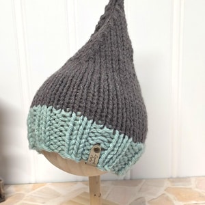 May include: A pointed knit hat featuring a dark gray upper section and a light mint green ribbed brim. A small, rectangular tan leather tag is attached to the brim, embossed with the text Laurel Arts in a dark brown script font.