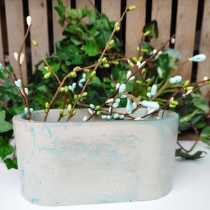 May include: A light gray concrete planter with a blue marbled design. The planter is oval shaped and is filled with a sprig of white and blue flowers.