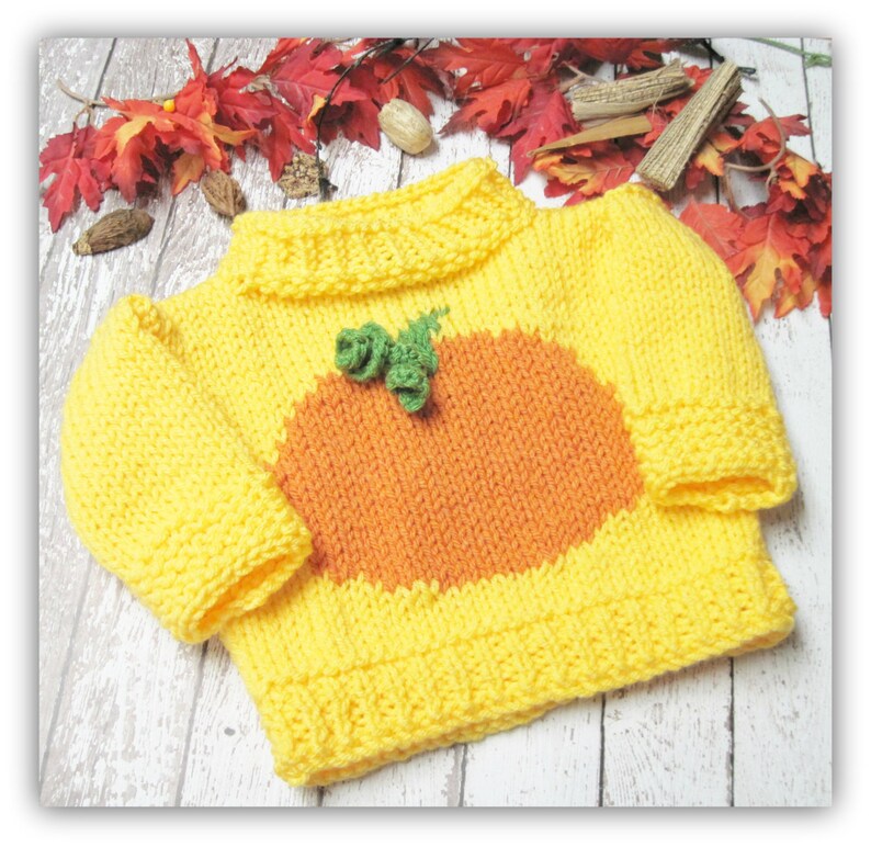 K&ouml;nnte beinhalten: Ein gelber Strickpullover mit einem orangefarbenen K&uuml;rbisdesign und gr&uuml;nen Bl&auml;ttern. Der Pullover hat einen Rundhalsausschnitt und lange &Auml;rmel.