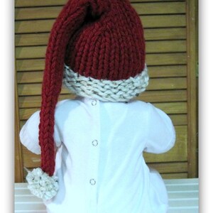 May include: A red knitted hat with a long, white pom-pom on the end. The hat has a white brim and is worn by a baby.