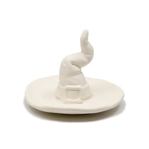 May include: A white ceramic witch hat ring holder. The hat has a curved, pointed top and a buckle detail on the brim. The ring holder is a Halloween-themed decorative item.