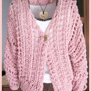 Könnte beinhalten: Hellrosa Strickjacke mit offenem Strickmuster, langen Ärmeln und einem einzelnen Knopfverschluss. Die Strickjacke wird auf einer Schaufensterpuppe präsentiert, darunter ein weißes Unterhemd und eine Holzanhänger-Halskette. Das Strickmuster hat ein strukturiertes Design.