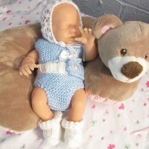 Puede incluir: Una muñeca bebé vestida con un conjunto de punto azul claro con detalles blancos, que incluye un gorro, un top, una braguita y patucos. Un lazo blanco y una perla adornan el conjunto. Un osito de peluche marrón está al lado de la muñeca.