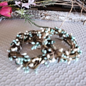 May include: Two small, round wreaths made of brown twigs, adorned with white and light blue berry-like accents. The wreaths are arranged on a textured, light-colored surface, suggesting a rustic or natural theme.
