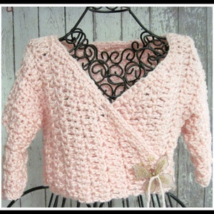May include: A pink crocheted wrap with a V-neck and long sleeves. The wrap is tied at the waist with a decorative bow.