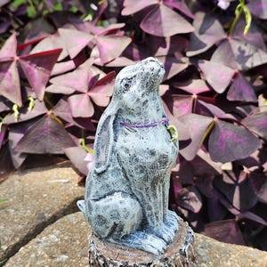 May include: A grey and white stone-textured rabbit figurine sitting upright on a circular wooden base with rough bark edges. The rabbit has a purple cord necklace featuring a small yellow crescent moon charm around its neck and looks upward.
