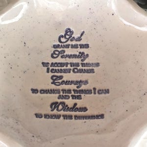 May include: A cream-colored ceramic dish with a scalloped edge. The dish features the text "God grant me the Serenity..." in dark blue script. The dish has a speckled appearance.