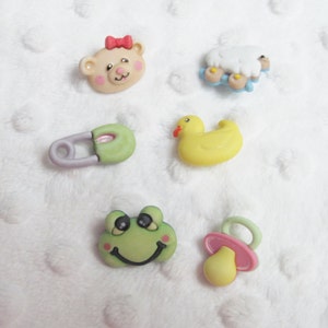 May include: Six colorful plastic buttons with baby-themed designs. The buttons feature a teddy bear, a lamb, a duck, a safety pin, a frog, and a pacifier.