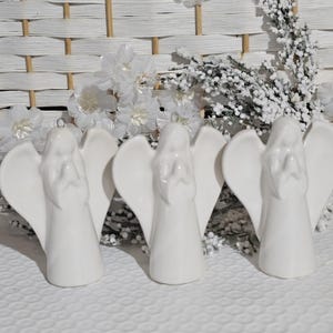 May include: Three white ceramic angel figurines with outstretched wings and clasped hands. The angels are standing on a white surface, with a white woven background and white floral accents. The angels are a religious or decorative item.