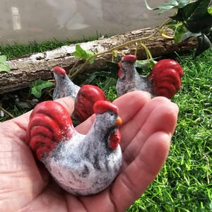 May include: Three small ceramic rooster figurines with white and gray bodies and red combs and tails. The roosters are sitting on a hand.