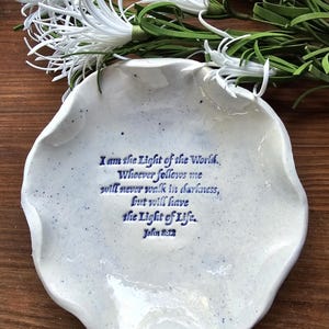 May include: White ceramic dish with a blue speckled glaze. The dish has a wavy edge and features a blue inscription with the text "I am the Light of the World. Whoever follows me will never walk in darkness, but will have the Light of Life. John 8:12"
