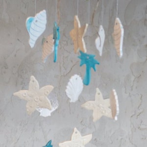 May include: A ceramic wind chime with a driftwood base and various sea-themed charms, including starfish, seashells, palm trees, and fish. The charms are painted in shades of blue, white, and beige.