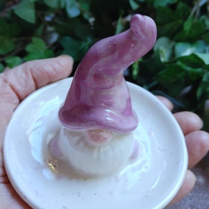 May include: A white ceramic ring dish with a purple gnome figurine. The gnome has a white beard and a pink nose. The dish is decorated with small white dots.