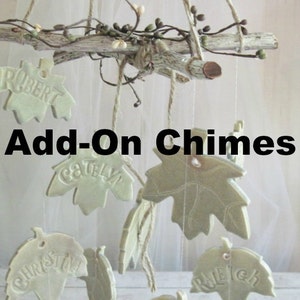 May include: A ceramic wind chime with leaf-shaped charms, each with a name inscribed on it. The names include Robert, Catlyn, Christina, Raleigh, and Frank. The chime is suspended from a branch with twine.