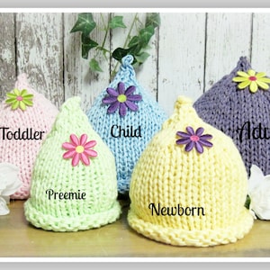May include: Five knitted hats in various colors, each adorned with a flower. The hats are labeled "Toddler," "Preemie," "Child," "Newborn," and "Adult." The hats are in shades of pink, green, blue, yellow, and purple. The hats are displayed on a wooden surface.