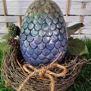 May include: A blue and green ceramic dragon egg with a scaly texture, sitting in a brown woven nest.
