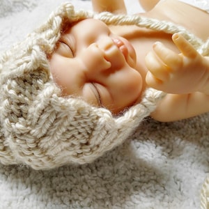 Op de afbeelding: Een pasgeboren baby met een beige gebreide muts met een textuurpatroon. De baby ligt op een witte, zachte ondergrond, met gesloten ogen en opgestoken vingers. De muts heeft een lange, gevlochten veter.