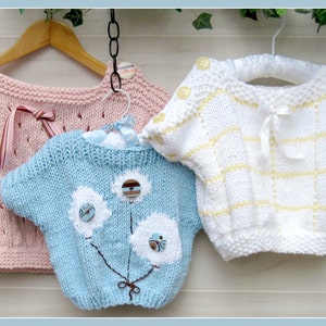 May include: Three knitted baby sweaters hanging on hangers. The sweaters are pink, blue, and white with different patterns and embellishments.