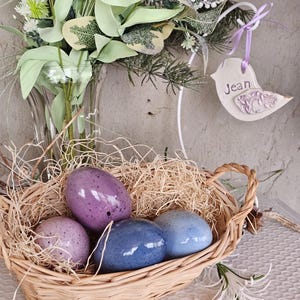 May include: A wicker basket filled with straw and four speckled, colorful eggs. The eggs are shades of purple and blue. A decorative bird-shaped tag with the name "Jean" hangs nearby. The background includes a vase of greenery.