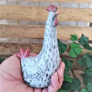 Puede incluir: Una figurita de cer&aacute;mica blanca de pollo con manchas negras y detalles rojos. El pollo est&aacute; de pie con la cabeza inclinada hacia un lado.