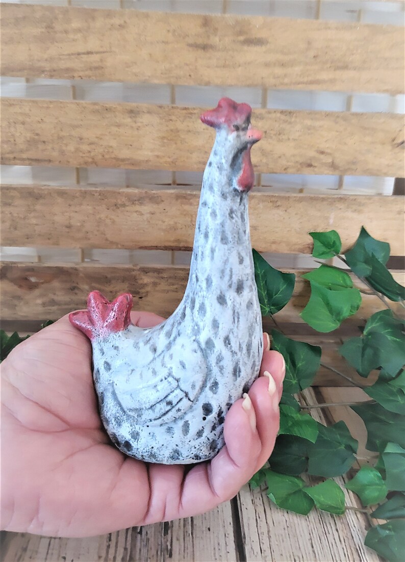 Puede incluir: Una figurita de cer&aacute;mica blanca de pollo con manchas negras y detalles rojos. El pollo est&aacute; de pie con la cabeza inclinada hacia un lado.