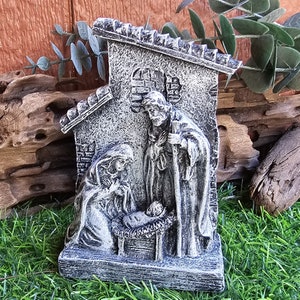 May include: A gray and white ceramic nativity scene with a small house, Mary, Joseph, and baby Jesus in a manger.