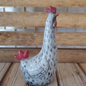 Puede incluir: Una figurita de pollo de cer&aacute;mica blanca con motas grises y detalles rojos. El pollo est&aacute; de pie con la cabeza inclinada hacia un lado.