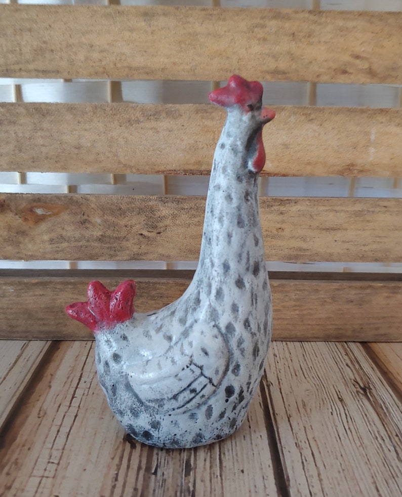 Puede incluir: Una figurita de pollo de cer&aacute;mica blanca con motas grises y detalles rojos. El pollo est&aacute; de pie con la cabeza inclinada hacia un lado.