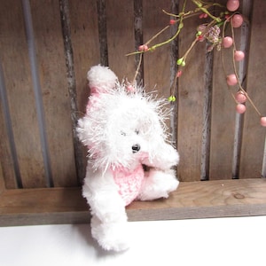 May include: A white plush toy dog wearing a pink knitted hat and a pink knitted outfit. The dog is sitting on a wooden surface.