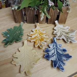 May include: Five ceramic leaf-shaped dishes in different colors. The dishes are green, yellow, white, blue, and cream. They are all in the shape of an oak leaf.