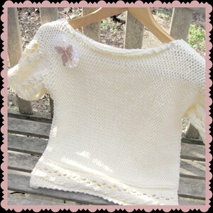 May include: A white knitted sweater with a delicate lace trim around the bottom and sleeves. A small, decorative butterfly brooch is pinned to the front of the sweater.