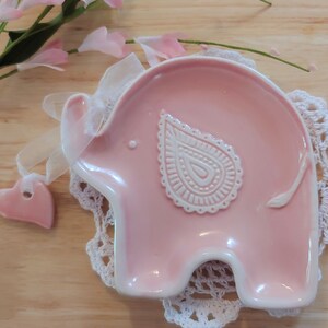May include: A pink ceramic elephant dish with a white paisley design. The dish is on a white crocheted doily.