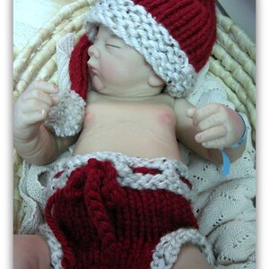 May include: A baby doll wearing a red and white knitted Santa hat and matching diaper. The doll is lying in a wicker basket.