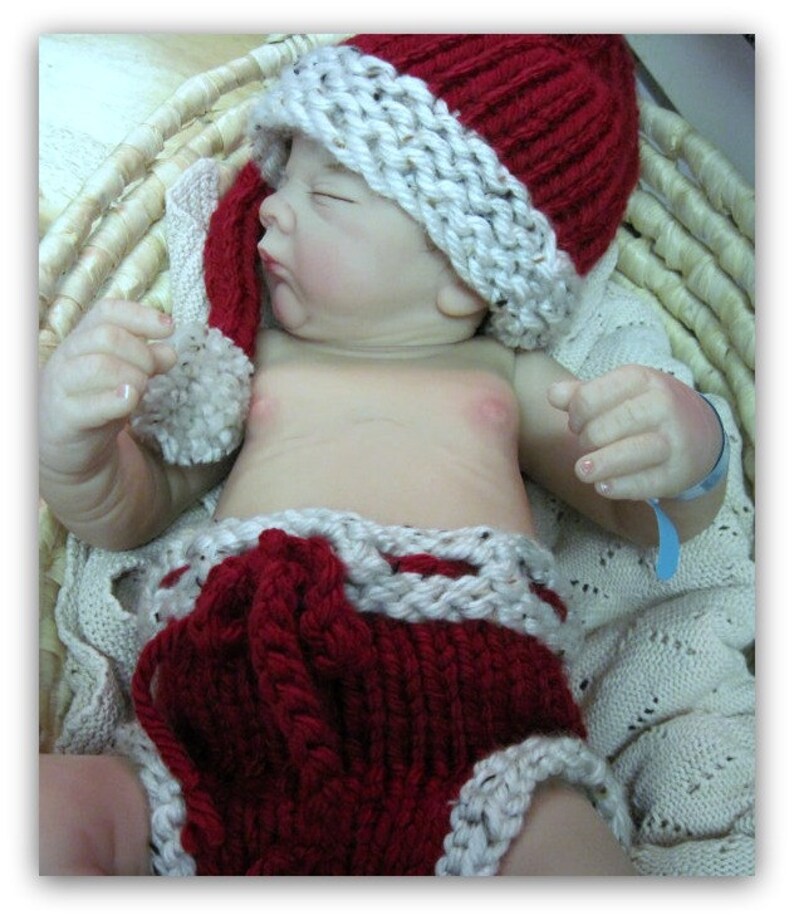 May include: A baby doll wearing a red and white knitted Santa hat and matching diaper. The doll is lying in a wicker basket.