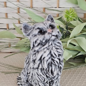 May include: A small, seated cat figurine with a black and white striped pattern. The cat has a pink nose and is set against a backdrop of greenery and a woven basket.