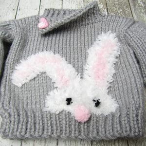 Puede incluir: Un suéter de punto gris con una aplicación de conejo blanco. El conejo tiene orejas rosas y una nariz rosa.