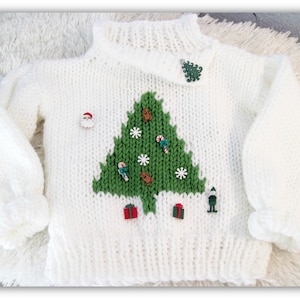 Könnte beinhalten: Weißer Strickpullover mit einem grünen Weihnachtsbaum-Design und verschiedenen festlichen Knöpfen, darunter ein Weihnachtsmann, ein Zuckerstange, ein Stern und ein Weihnachtsbaum.