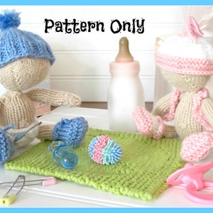 Puede incluir: Dos muñecas de punto, una en azul y otra en rosa, con gorros y botines a juego. Las muñecas están sentadas en una manta de punto verde con una botella, un chupete y una pelota de colores.