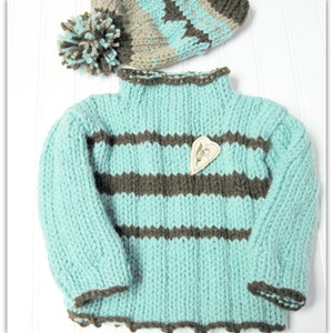 Puede incluir: Un suéter de punto a rayas azul claro y marrón con ribete marrón. El suéter tiene cuello alto y mangas largas. Una pequeña etiqueta en forma de corazón está prendida en la parte delantera del suéter.