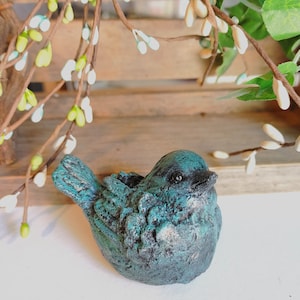 May include: A small, blue-green bird figurine with a distressed finish. The bird is perched on a wooden surface with white flowers in the background.