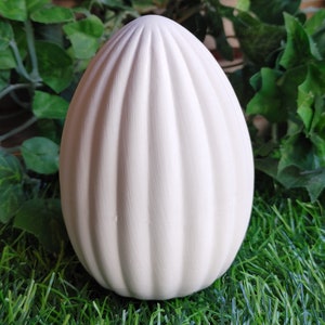 May include: White ceramic egg with a ribbed texture. The egg is sitting on a bed of green artificial grass.